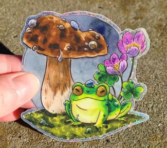 A glittery vinyl sticker of a little green frog happily sitting beneath a mushroom. There are raindrops on the frog, mushroom, and on the sorrel flowers behind.